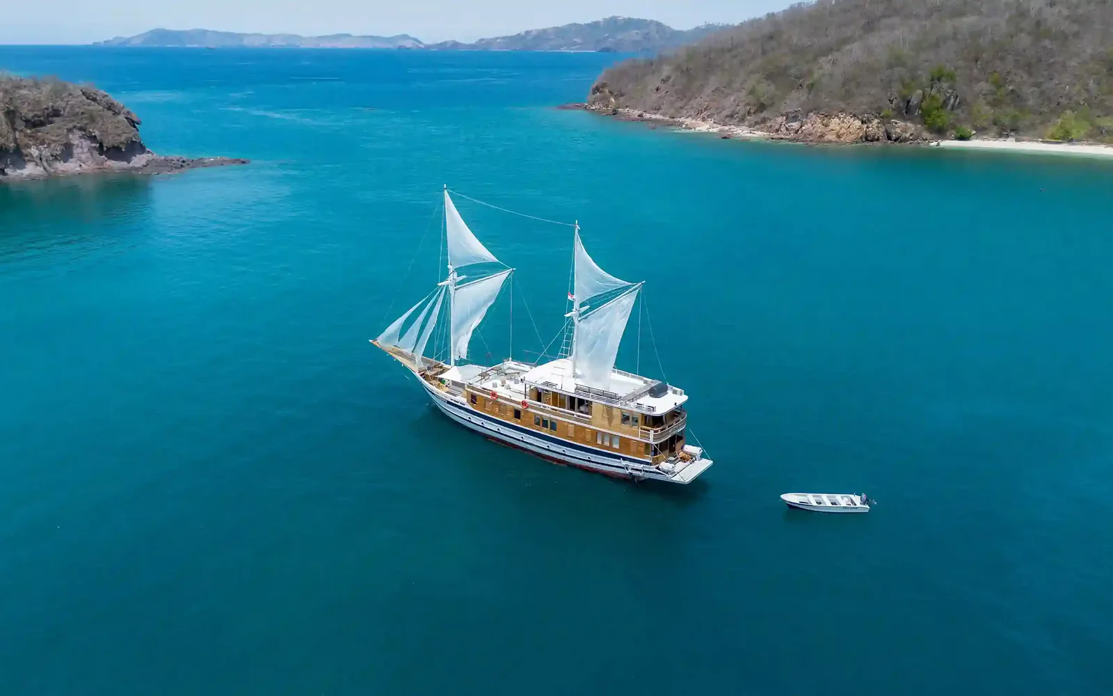 A phinisi lying at anchor under sail in a turquoise bay